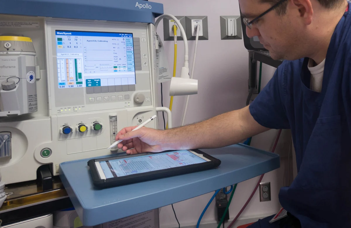 Clinician using AnesthesiaEMR on an iPad beside an anesthesia machine.